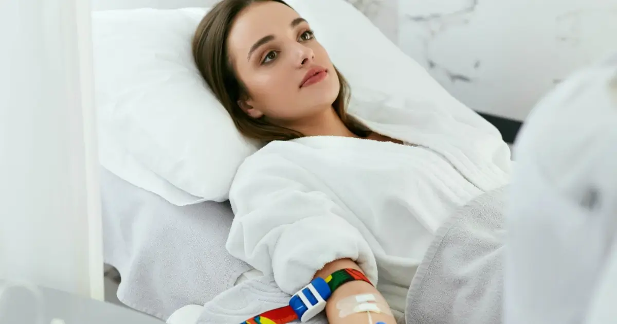 A woman in a white robe receives IV Ozone Therapy in Long Island, NY, from a clinician in blue gloves, with a medical monitor visible nearby.